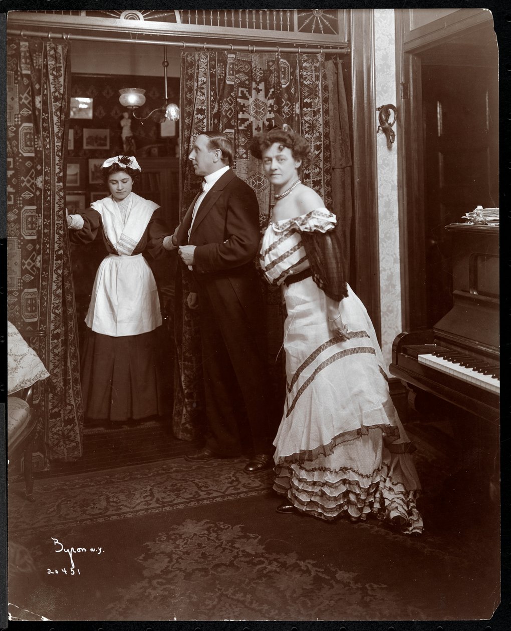 Ein formell gekleideter Mann und eine Frau sowie eine offenbar häusliche Dienerin in einem Raum mit einem Klavier, 1905 von Byron Company