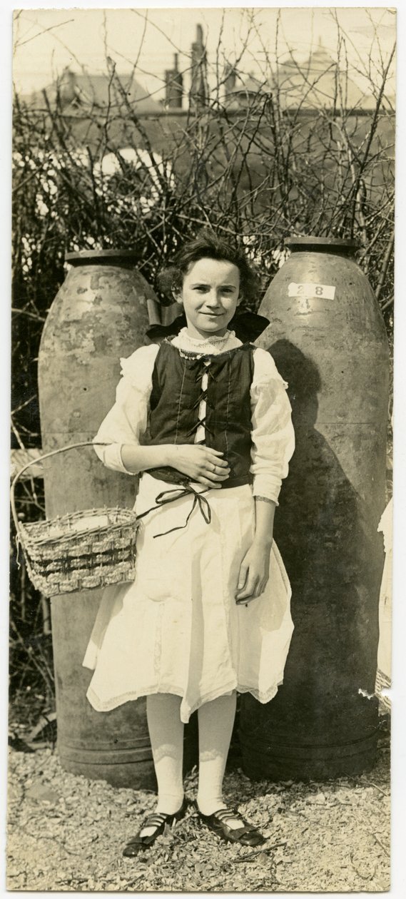 Vertikales, sepiafarbenes Foto eines Mädchens in einem Kleid vor zwei großen Artilleriegranaten. Das Mädchen hält einen Korb in der Armbeuge ihres rechten Arms von Carl Michel